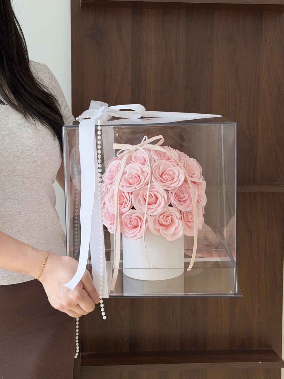 A person holds a clear acrylic gift box containing a bouquet of pink roses in a white box, with a white ribbon and bow on a dark wood shelf background.