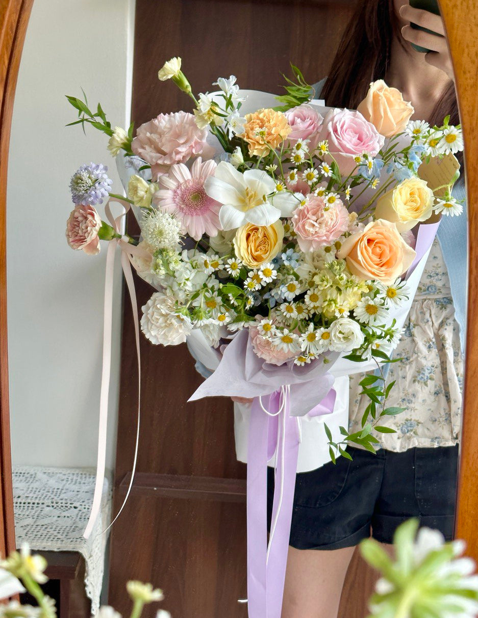 Person holding a bouquet of flowers mixed with champagne rose, buttercup rose, white tulip, pink gerbera, chamomile, delphinium blue and other fillers in front of a mirror.