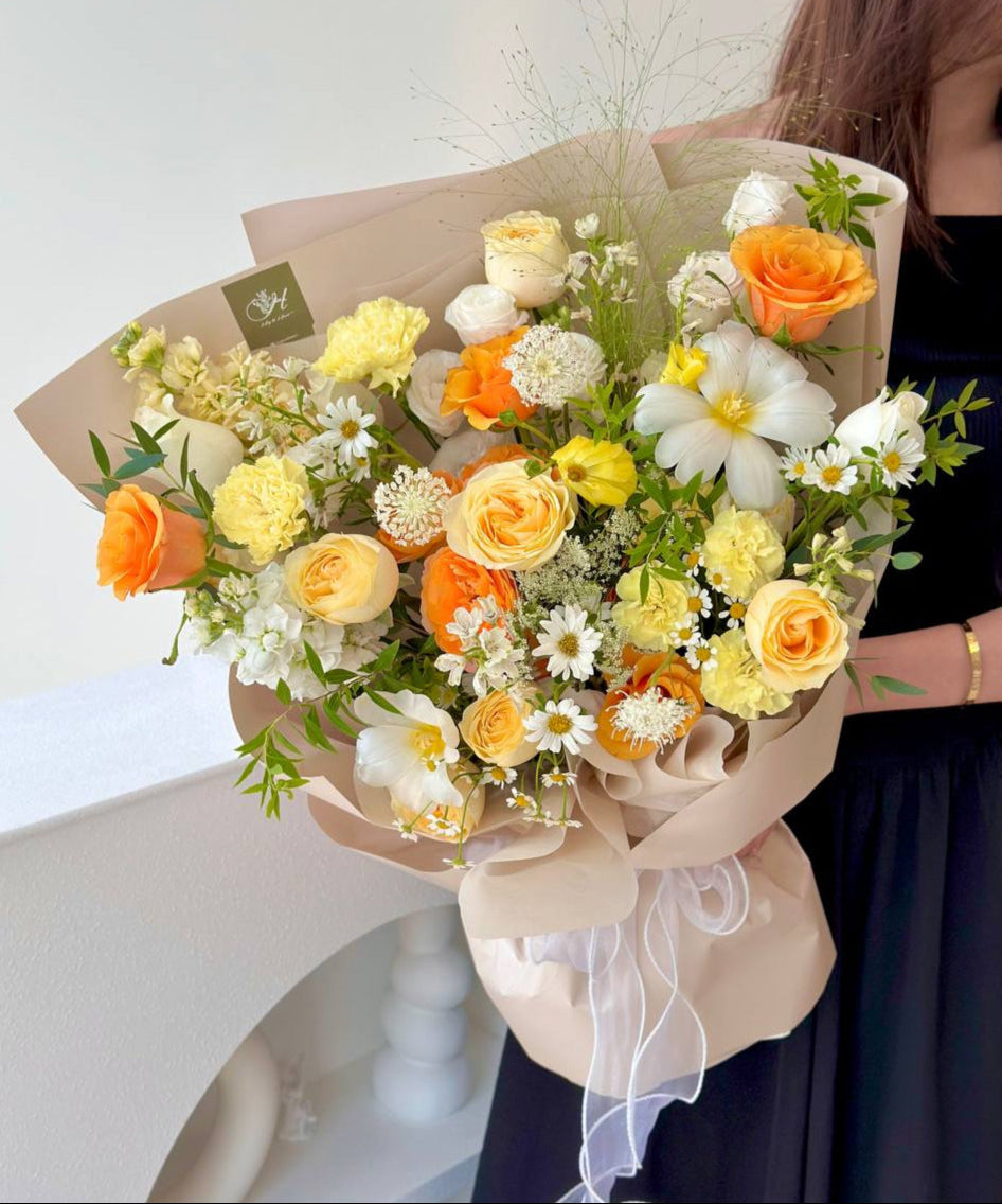 A cheerful bouquet featuring yellow, orange, and white blooms, wrapped in soft beige paper with a white ribbon.

