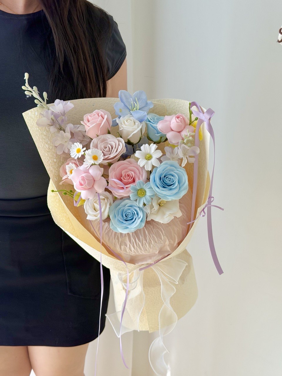 A person holding a pastel flower bouquet with pink, light blue, and white roses wrapped in cream paper with purple ribbon ties, against a plain light background.