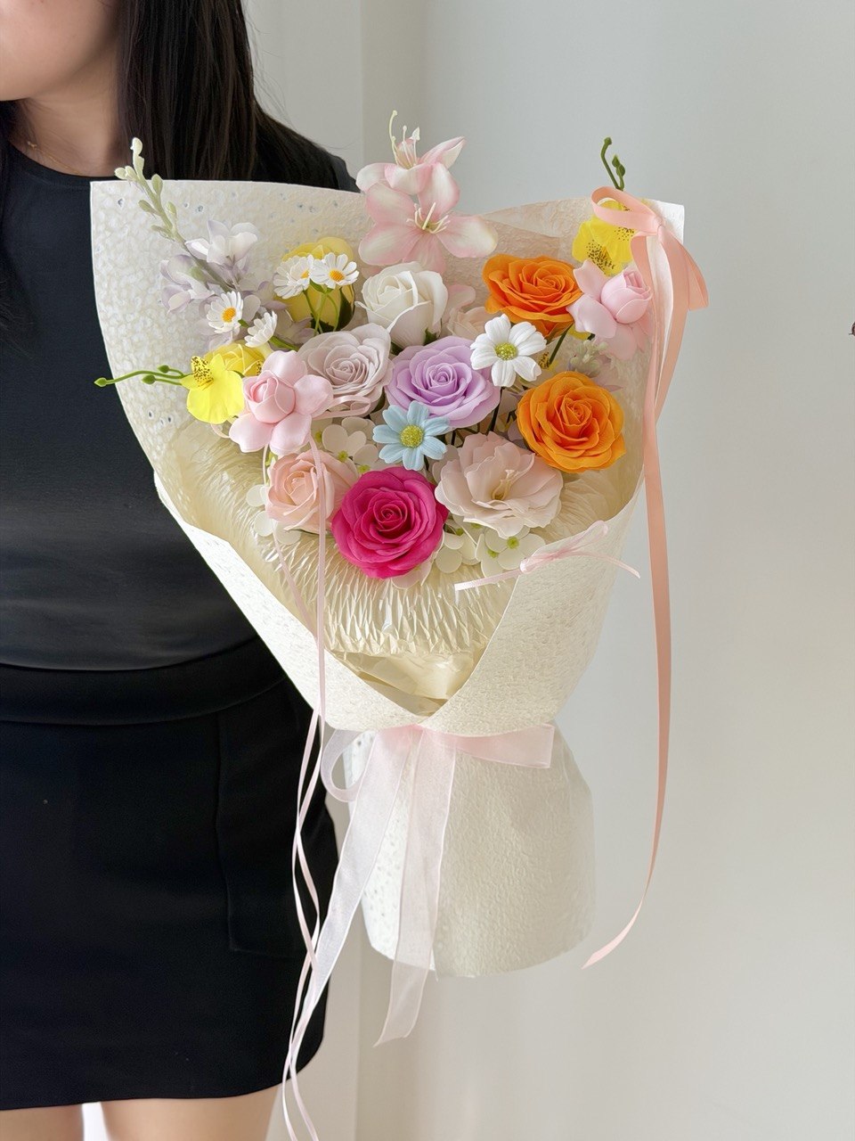 A person holding a colorful flower bouquet wrapped in cream paper with a pale pink ribbon. The bouquet contains pink, orange, yellow, purple, and white flowers.