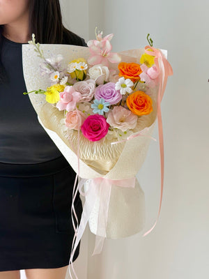 A person holding a colorful flower bouquet wrapped in cream paper with a pale pink ribbon. The bouquet contains pink, orange, yellow, purple, and white flowers.
