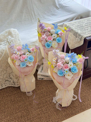 Three pastel soap flower bouquets wrapped in cream paper, arranged on a carpeted floor in front of a white draped sofa. The bouquets contain pink, blue, and white artificial roses with small yellow and purple filler flowers, tied with lavender ribbons.