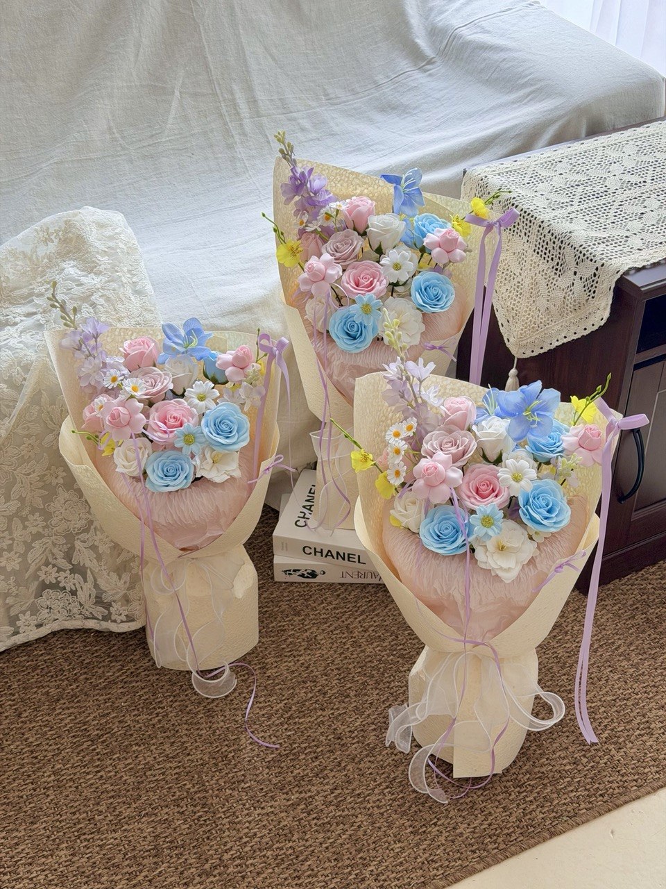 Three pastel soap flower bouquets wrapped in cream paper, arranged on a carpeted floor in front of a white draped sofa. The bouquets contain pink, blue, and white artificial roses with small yellow and purple filler flowers, tied with lavender ribbons.