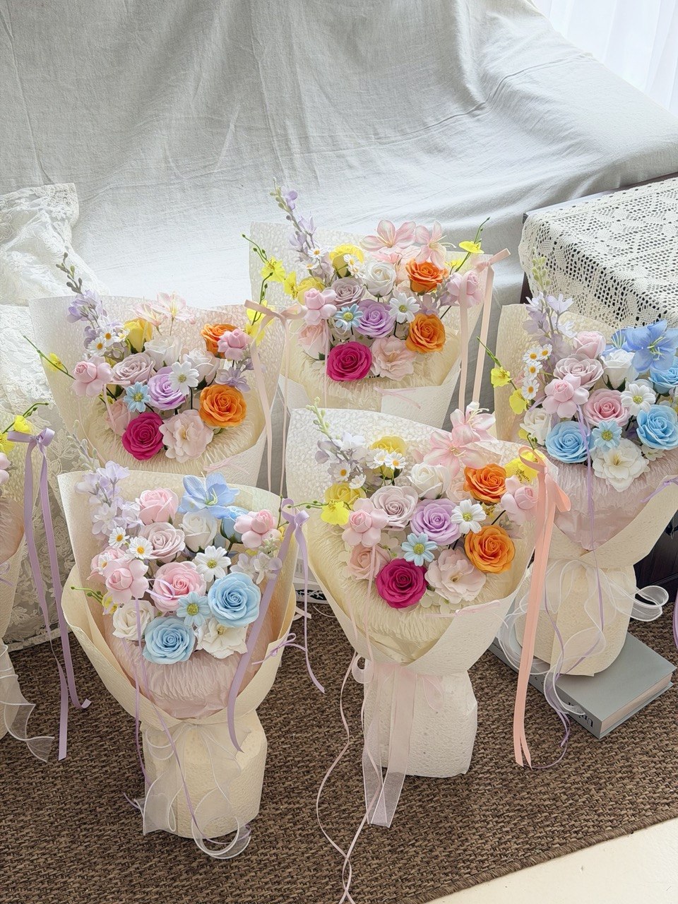 Five pastel-colored soap flower bouquets wrapped in cream paper and tied with ribbons, arranged on a carpeted floor against a white fabric backdrop.