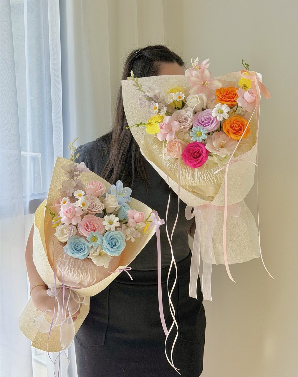 A person wearing a black top holds two large, colorful soap flower bouquets wrapped in cream paper. The bouquets feature artificial roses in pastel pink, blue, orange, purple, and white, with small daisy-like flowers and sheer ribbon ties.
