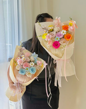 A person wearing a black top holds two large, colorful soap flower bouquets wrapped in cream paper. The bouquets feature artificial roses in pastel pink, blue, orange, purple, and white, with small daisy-like flowers and sheer ribbon ties.