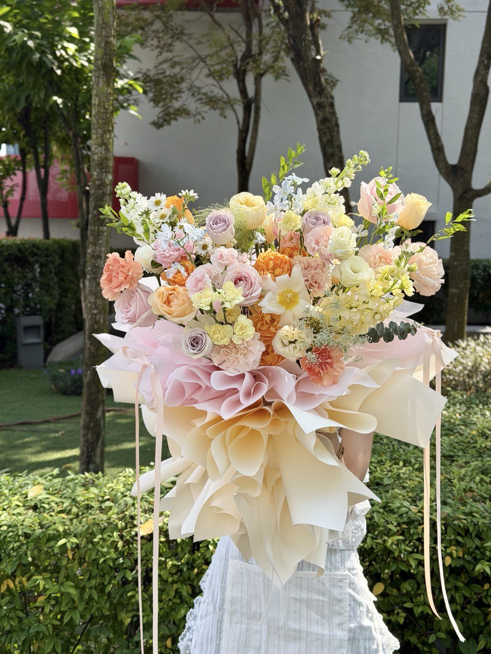 A girl wearing a white dress holding a mixed colour roses with white tulip, pink rose, lilac rose, delphinium blue and fillers witha ballet wrapping at hug and heart florist.