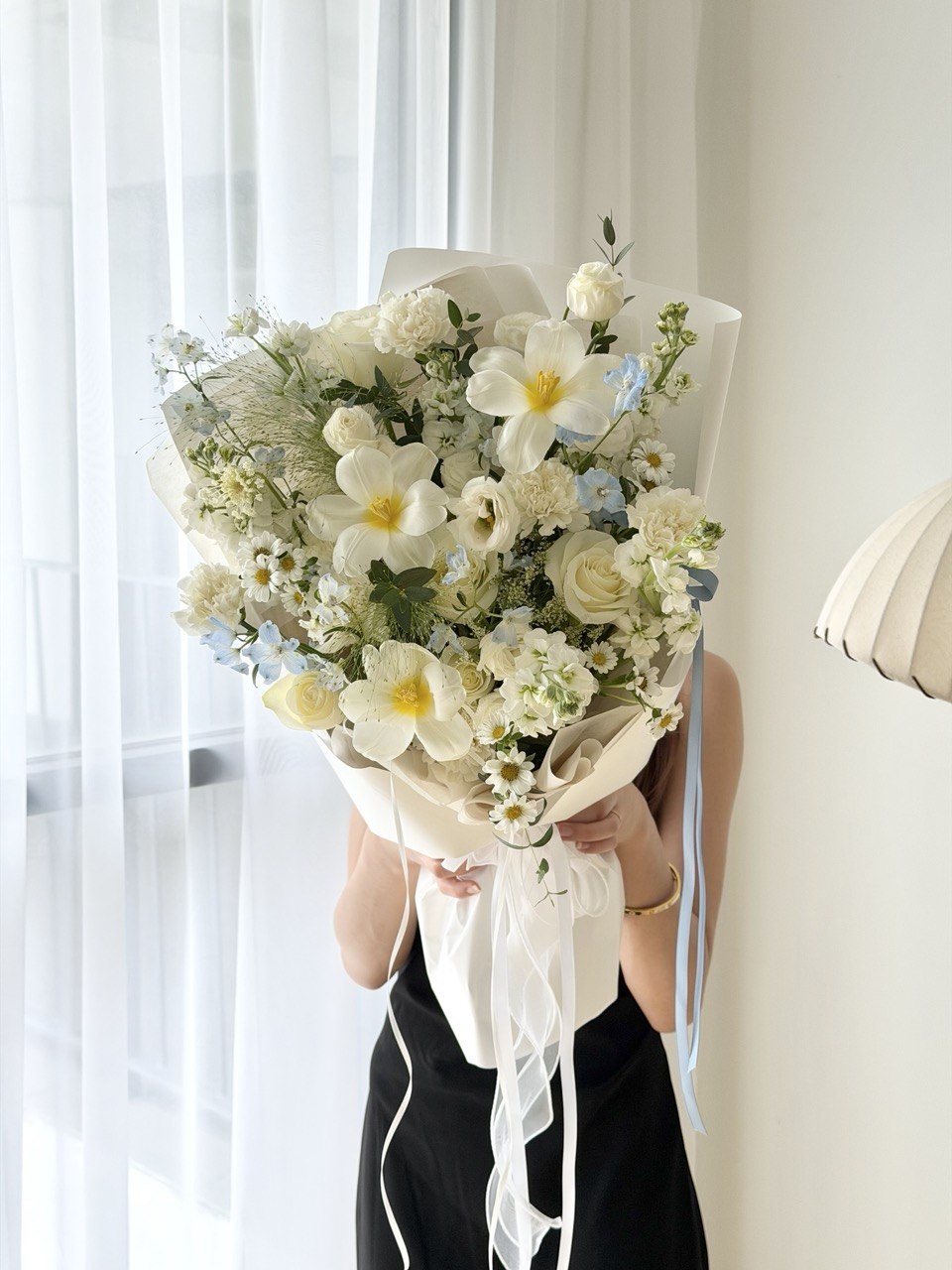 Premium white flower bouquet with soft blue accents, white roses, carnations, daisies and seasonal flowers, beautifully held by a girl for special occasions.