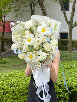 Elegant white and light blue flower bouquet with white roses, white plumeria, blue delphinium, daisies and carnations, held by a girl for birthday or anniversary gift.