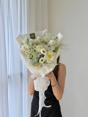 A person in a black dress holds a large bouquet of white and light blue flowers wrapped in cream paper, standing in front of a white curtain and wall.