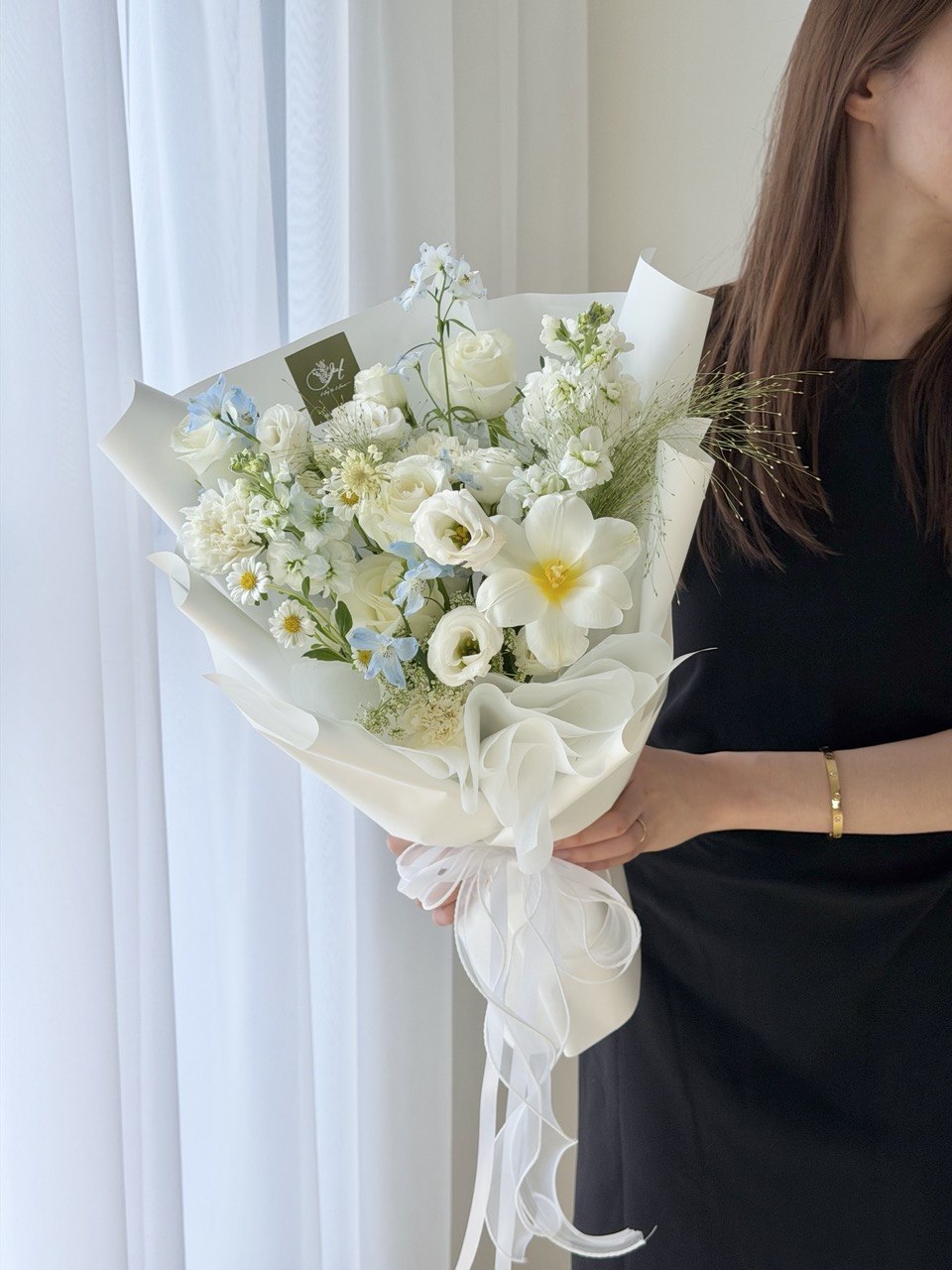 A person in a black dress holds a bouquet of white and light blue flowers wrapped in white paper with a sheer white ribbon, set against a soft white curtain background.