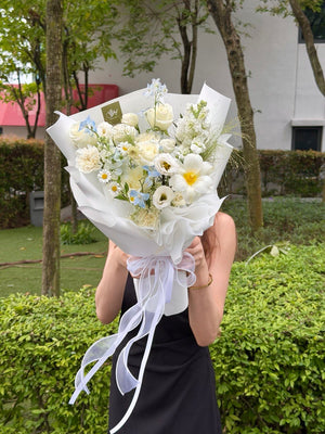 A person outdoors holding a large bouquet of white and light blue flowers wrapped in white paper with a white ribbon, partially obscuring their face. The background shows green hedges and trees.