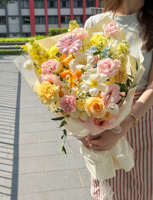 A person outdoors holding a large, vibrant bouquet of assorted pastel flowers, including pink, yellow, peach, and white blooms, wrapped in cream paper.