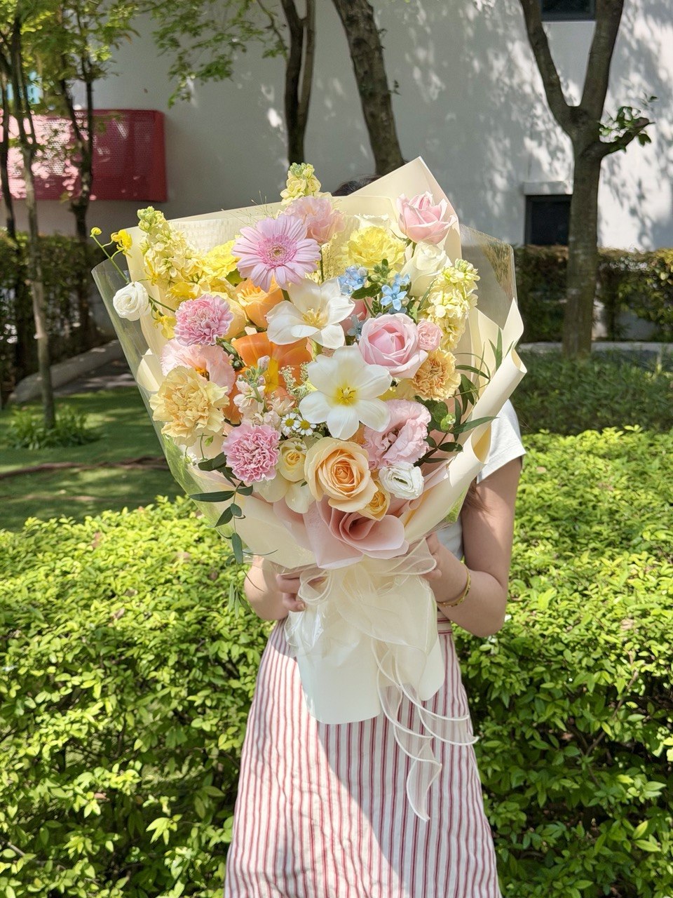 A person outdoors holding a large pastel flower bouquet with pink, yellow, peach, and white blooms wrapped in cream paper. The background shows green hedges and trees.