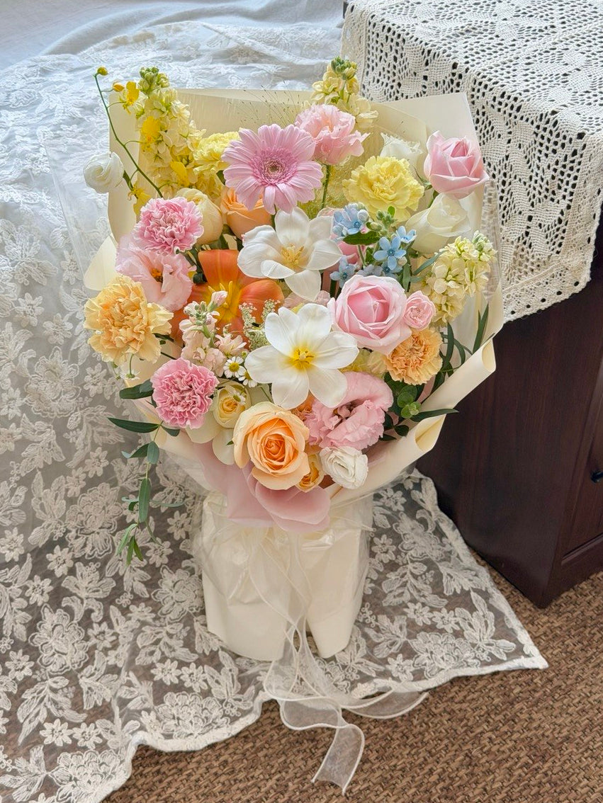 A pastel flower bouquet wrapped in cream paper, featuring pink, yellow, white, and peach blossoms with green foliage, placed on a lace-covered surface next to a dark wooden table.