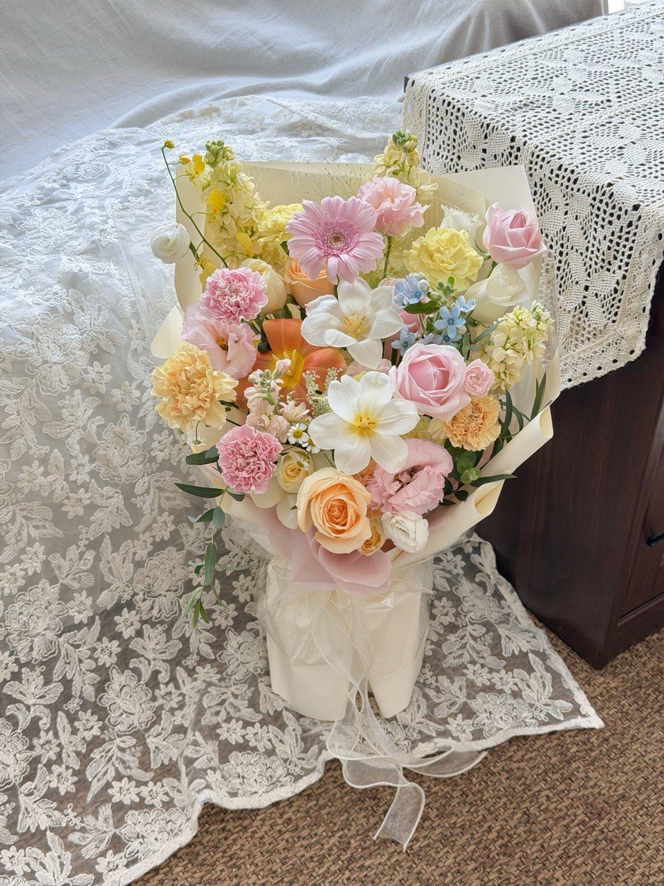 A pastel flower bouquet wrapped in cream paper, featuring pink, yellow, white, and peach blossoms with green foliage, placed on a lace-covered surface next to a dark wooden table.