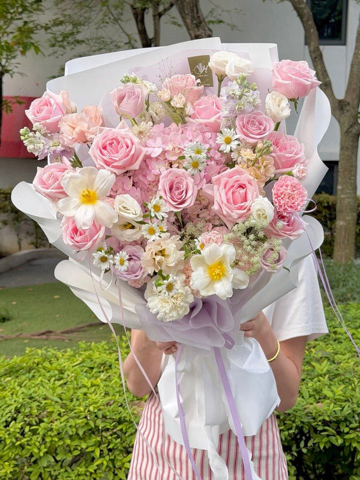 A person outdoors holding a large bouquet of pink roses, pink hydrangea and white flowers wrapped in white paper with a lavender ribbon. The background shows green hedges and trees.