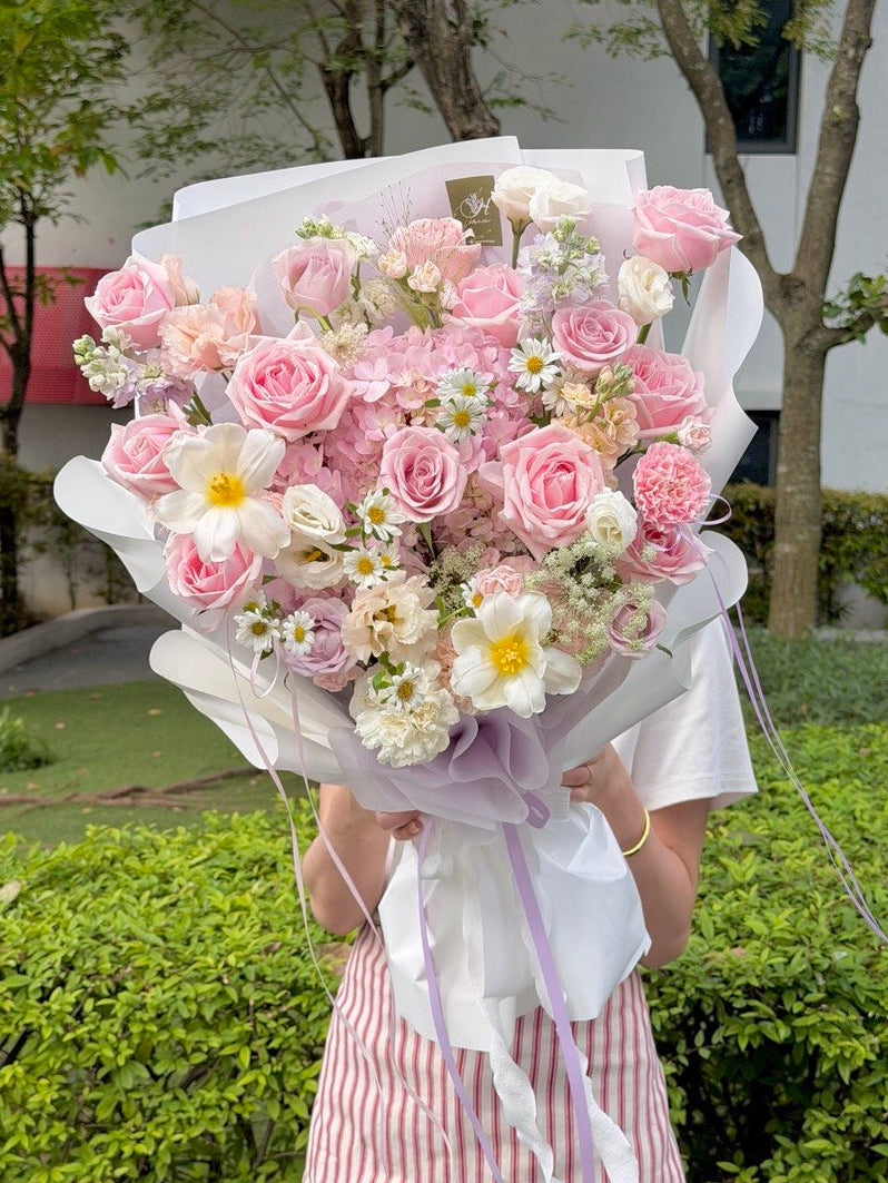 A person outdoors holding a large bouquet of pink roses, pink hydrangea and white flowers wrapped in white paper with a lavender ribbon. The background shows green hedges and trees.