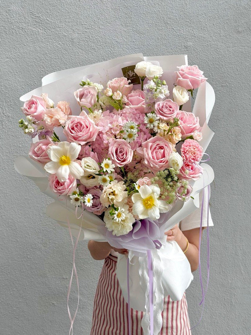 A large bouquet of pink roses, pink hydrangea and white flowers wrapped in white paper with purple ribbon accents, held by a person wearing a pink and white striped dress against a textured gray wall.
