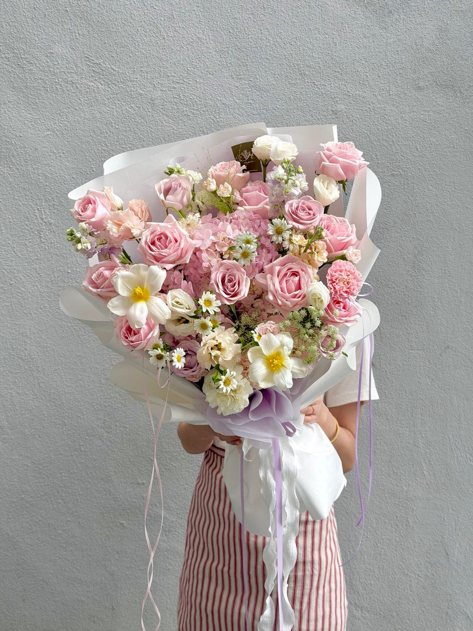 A large bouquet of pink roses, pink hydrangea and white flowers wrapped in white paper with purple ribbon accents, held by a person wearing a pink and white striped dress against a textured gray wall.