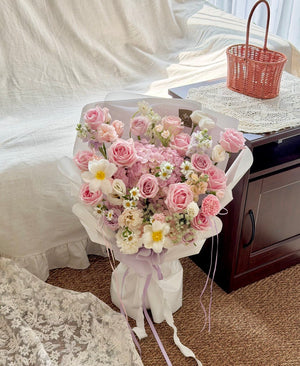 A large bouquet of pink hydrangea, pink roses and white flowers wrapped in white paper with a pale purple ribbon, placed on a carpeted floor next to a bed and a small woven basket.