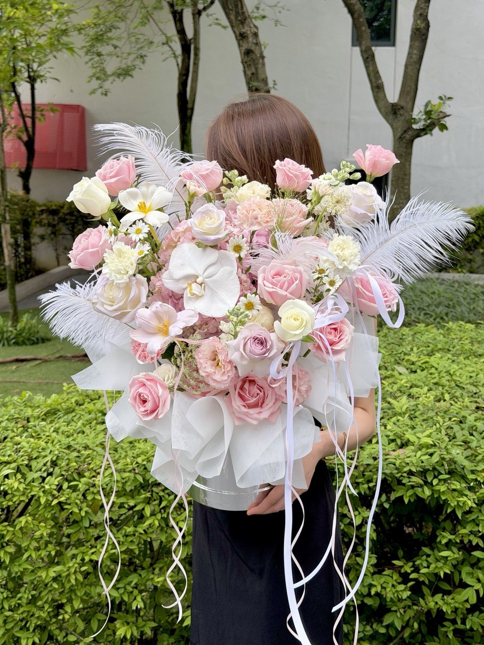 A person outdoors holding a large, elaborate floral bouquet. The bouquet features pink and white roses, Pink Hydrangea, white orchids, white daisies, white feathers, and white ribbon streamers wrapped in sheer white paper.