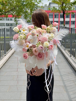 A woman stands on an outdoor walkway holding a large bouquet of pink and white flowers with white feathers and long white ribbons. The photo is taken from behind, showing her from the back against a city background with trees and buildings.