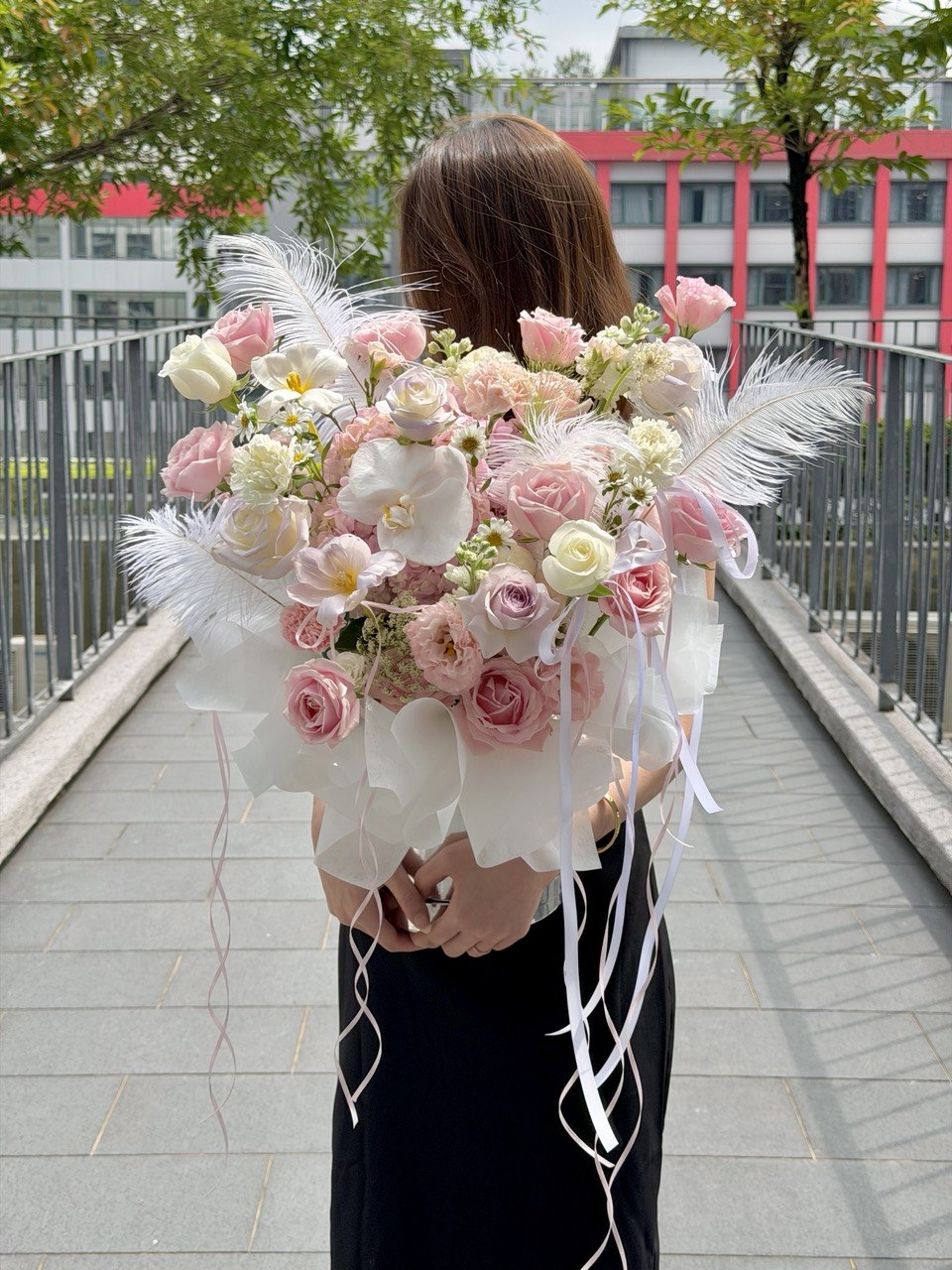 A woman stands on an outdoor walkway holding a large bouquet of pink and white flowers with white feathers and long white ribbons. The photo is taken from behind, showing her from the back against a city background with trees and buildings.