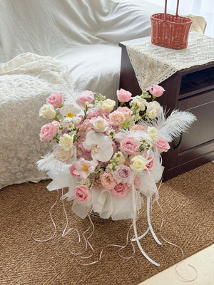 A large, elaborate floral arrangement box, featuring pink and white roses, orchids, daisies, pink hydrangea, white feathers, and white ribbon drapes, placed on a brown carpeted floor next to a bed and bedside table.