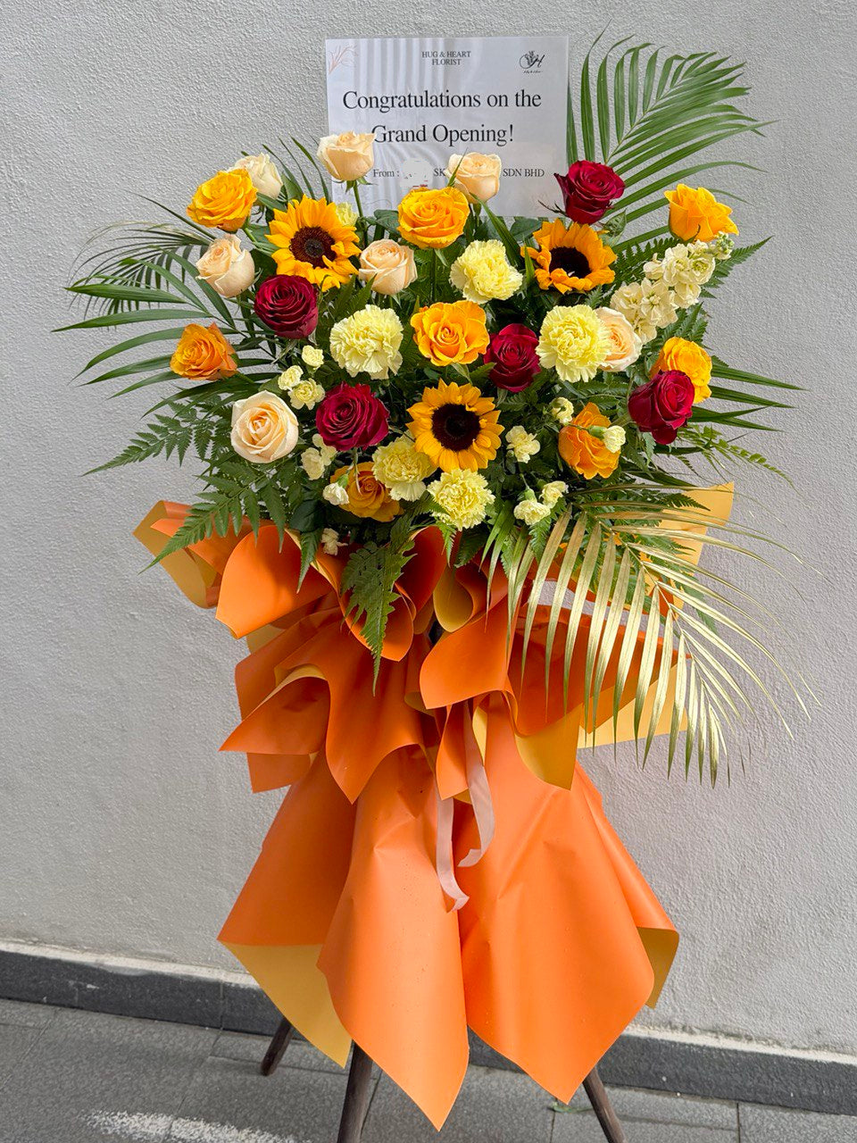 Floral arrangement with orange ribbon and 'Congratulations on the Grand Opening!' card against a gray background perfect for grand opening flower stand gift prepared by hug and heart florist at Sunway Geo.