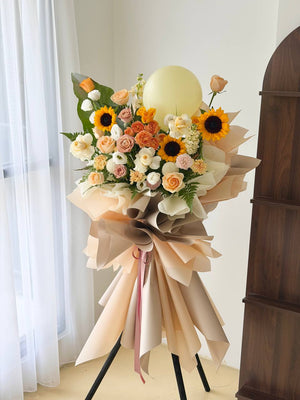 A large grand opening flower stand, featuring sunflowers, roses, and daisies in peach, orange, cream, and yellow tones, with a pale yellow balloon floating above and beige paper wrapping around the bouquet.