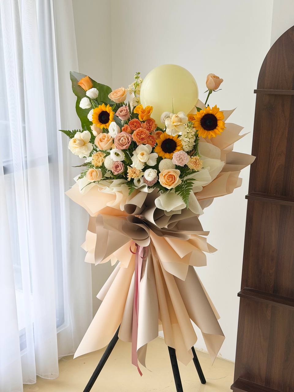 A large grand opening flower stand, featuring sunflowers, roses, and daisies in peach, orange, cream, and yellow tones, with a pale yellow balloon floating above and beige paper wrapping around the bouquet.