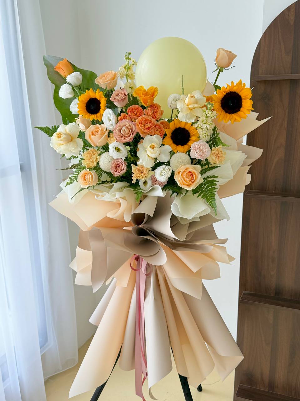 A large grand opening flower stand with a bouquet of sunflowers, roses and other pastel flowers, wrapped in beige paper with a large bow and ribbon, topped with a pale yellow balloon, photographed indoors near a window and wooden cabinet.