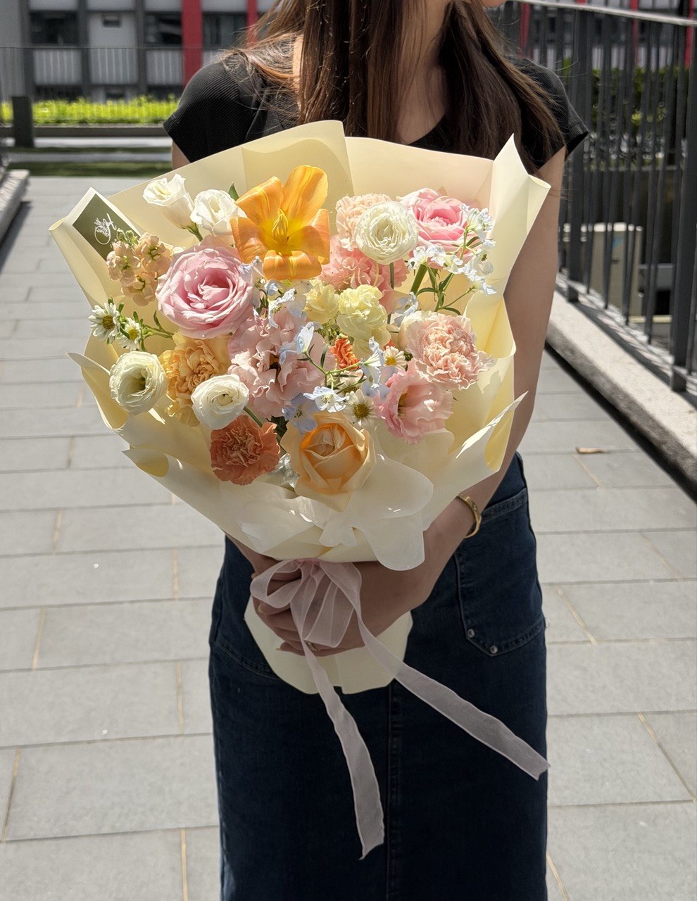 A person outdoors holding a small bouquet of pastel flowers wrapped in cream paper with a pale pink ribbon. The bouquet includes pink, peach, yellow, and white blossoms.