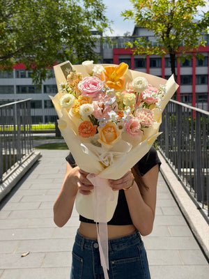 A person outdoors holding a small bouquet of pastel flowers wrapped in cream paper. The bouquet contains pink, peach, orange, and white flowers with a light pink ribbon.
